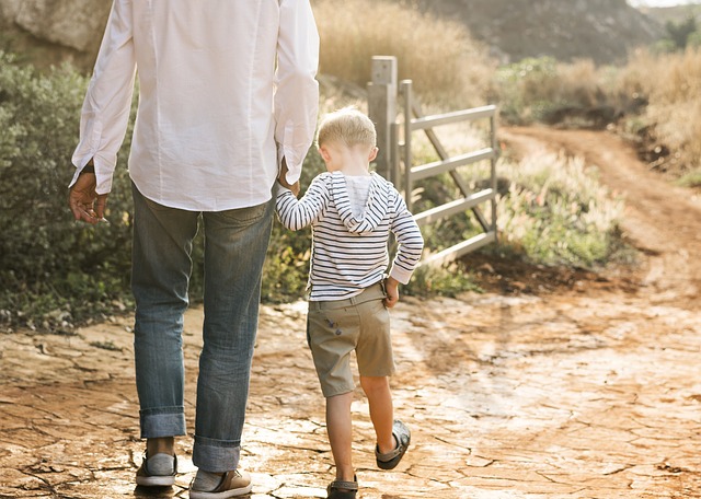 A young boy walking outside, holding his father's hand. They are facing away from the camera.