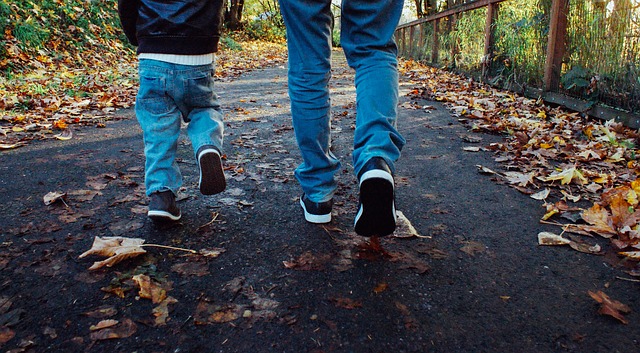 A young boy and his father walking down a road in fall. They are both wearing jeans and sneakers and walking away from the camera.