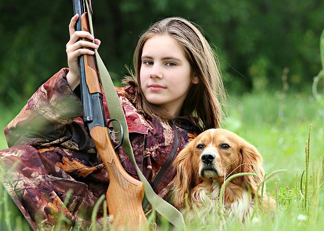 A young girl, wearing camo and sitting in a field, with a rifle and a dog.