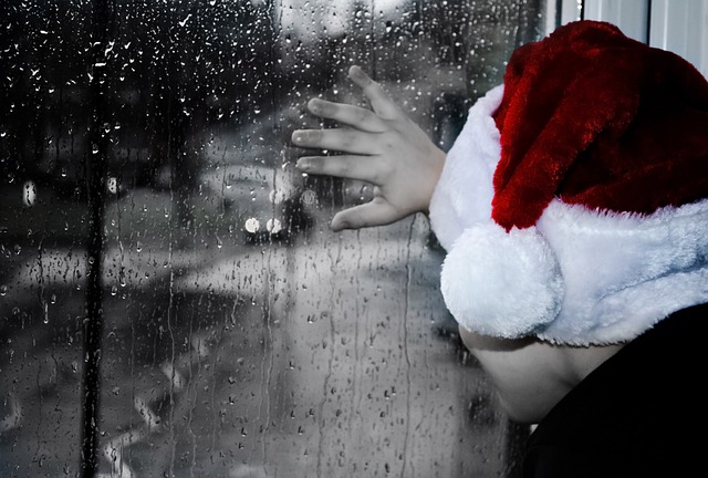 A child wearing a Santa hat looking out of a window onto a grey, rainy day and looking sad.