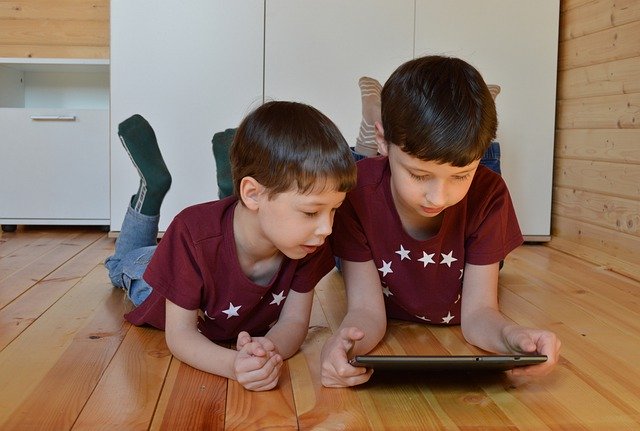 Two little boys who are clearly brothers, lying on the floor side by side and looking at a tablet screen together.