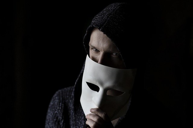 A man against a dark background, holding up a mask that partially covers his face.