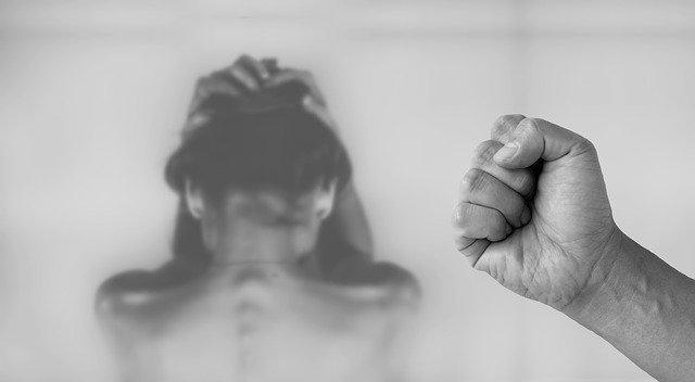 A black and white image of a woman with her face turned away from the camera, holding her head. A man's fist is in shape focus in front of her.