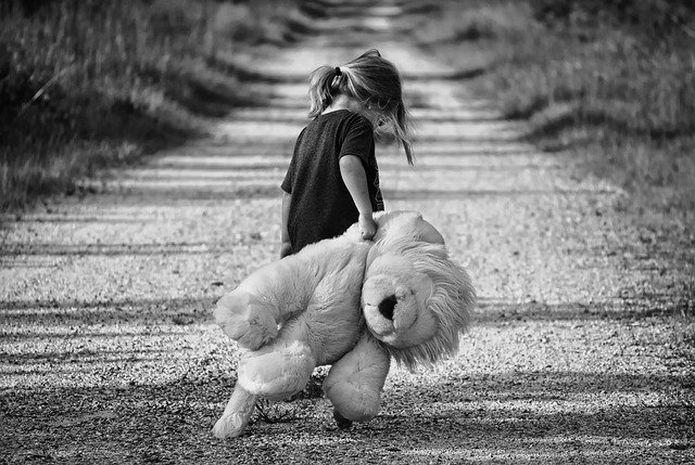 A black and white picture of a little girl, holding a large teddy bear and looking sad, walking away from the camera down a dirt road.